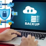 Persona usando una computadora portátil que muestra en pantalla un gráfico de respaldo digital con icono de nube, flechas de subida y bajada, y una base de datos. A la izquierda, hay ilustraciones de ciberseguridad como un candado, escudo y elementos de red, representando protección de datos y respaldo DIGITAL